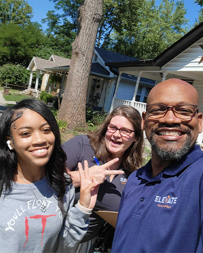 Three people smiling and waving at the camera in front of a home.