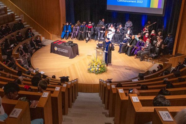 A speaker stands at a podium on stage in front of an audience. A group of people is seated on stage behind the speaker.