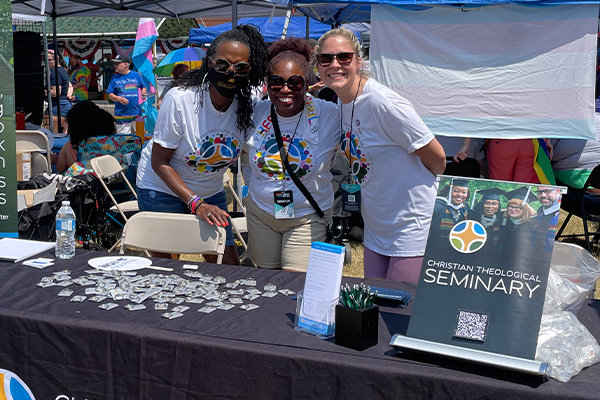 Three women, one in a mask, smile for a picture while standing arm in arm behind a table with small trinkets on display. The table displays a small pop-up sign that says "Christian Theological Seminary."
