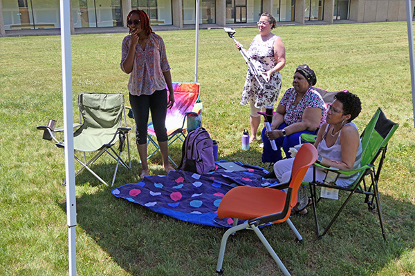 Two women standing and two women seated in camp chairs under a tent face forward in a reflective manner.