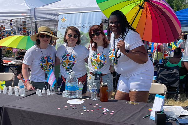 Four women, on holding a rainbow umbrella, stand arm in arm behind a table with water bottles, small containers, and small buttons on display.