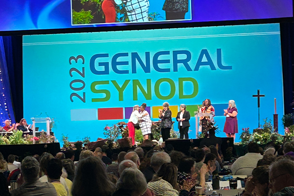 A crowd applauds facing a stage as a two women hug on stage while four additional women applaud on stage. There is a screen behind the stage that reads "2023 General Synod."