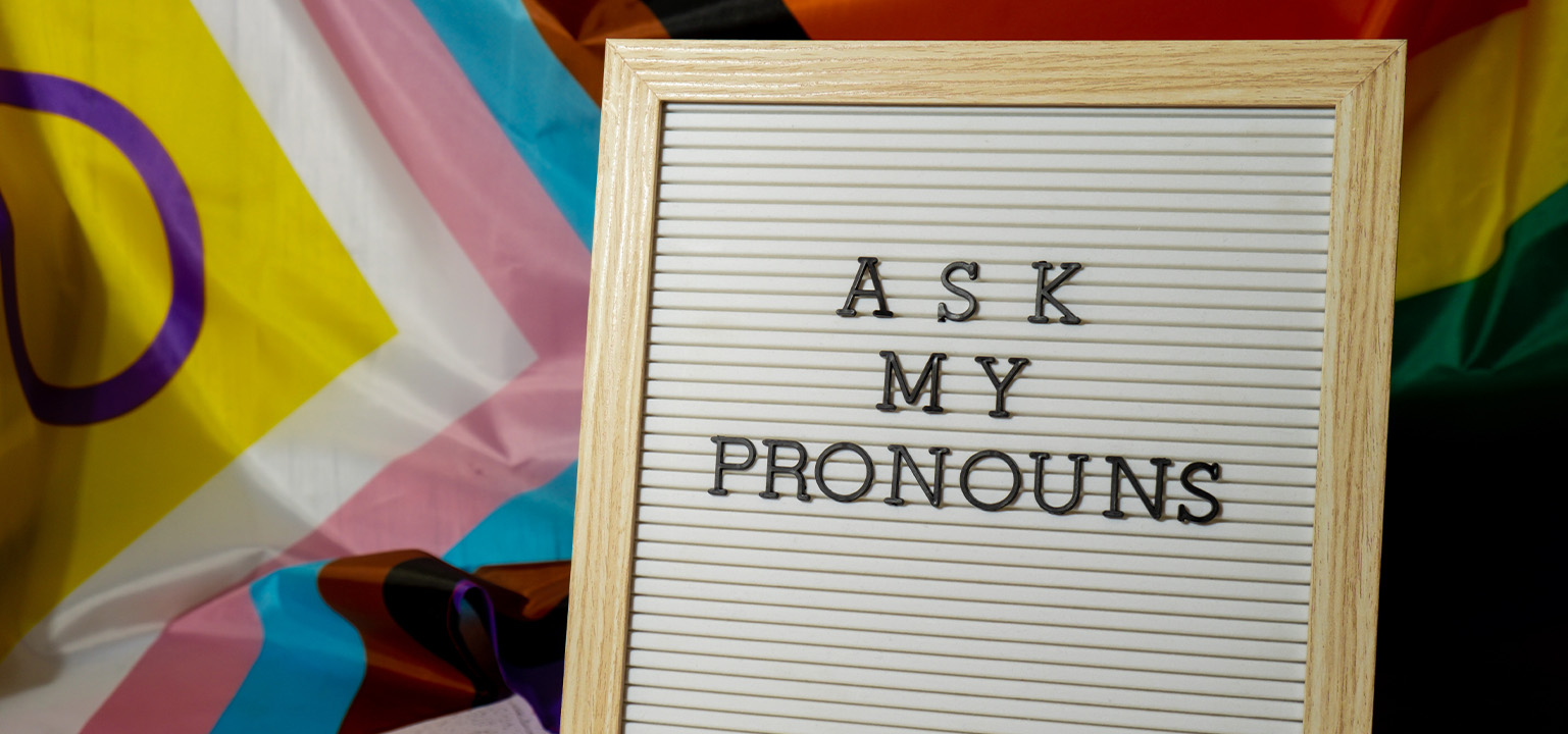 A board in front of the LGBTQ+ flag, with "Ask my pronouns" written on it.