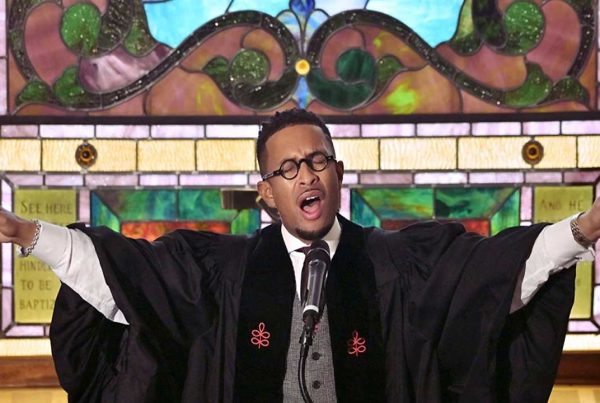 African American man delivers a sermon in front of a stained glass window.