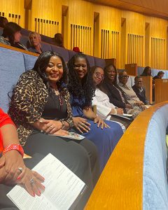 Six people smile at the camera and sit side by side on blue padded seating in a round theater with wooden walls.