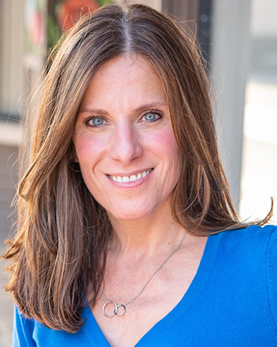 Andrea Zacharakis A woman with brown hair smiles looking at the camera. She is wearing a blue shirt.